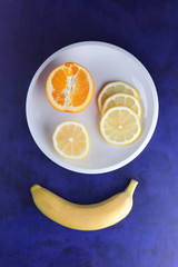 Citrus on ultraviolet background in minimalism style, slices of lemon and half orange on white plate, yellow banana on dark blue background, healthy food, vegan, pop art
