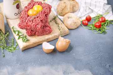 Fresh beef meat in forcemeat, on a wooden board. Raw eggs and tomatoes are small. Herbs for cooking and round buns. On a gray stone background. Free space for writing text.