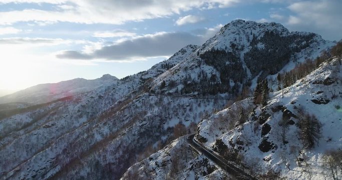Forward aerial top view over car travelling on road in Winter snow mountain near forest woods.Snowy mountains street path establisher.Sunset or sunrise sun backlit.4k drone flight  establishing shot
