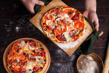 Close up hands of chef baker holding a board with hot freshly baked pizza at kitchen, view from above. Italian restaurant concept