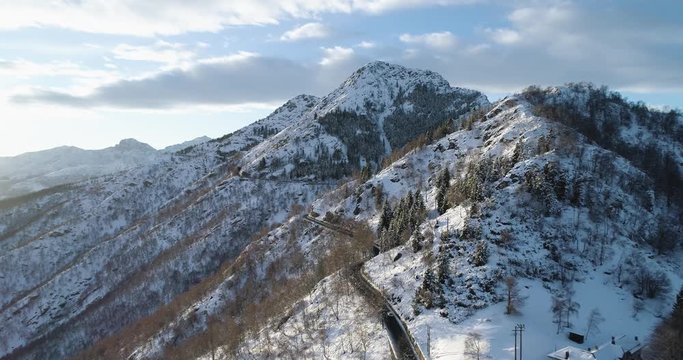 Forward aerial top view over car travelling on road in Winter snow mountain near forest woods.Snowy mountains street path establisher.Sunset or sunrise sun backlit.4k drone flight  establishing shot