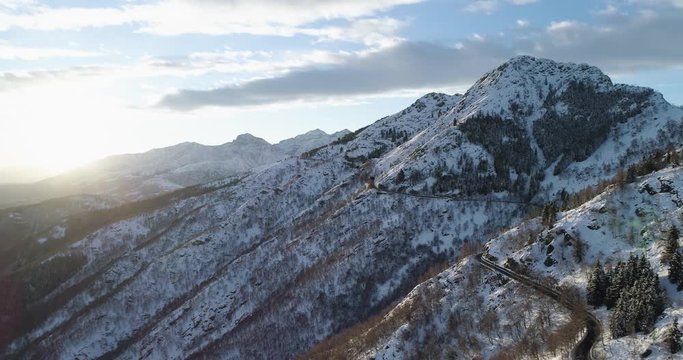 Forward aerial top view along road on white snow mountain in winter.Forest woods.Snowy mountains street path establisher.Sunset or sunrise sun backlit.4k drone flight straight-down establishing shot