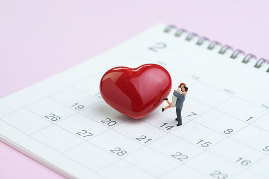 Miniature figures sweet couple standing with shiny red heart shape on 14th February calendar on pink background as Valentine's day concept.