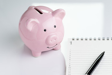 Personal finance to do lists or new year's resolution for savings concept, pink piggy bank and white notepad with numbers listed and pen on white table
