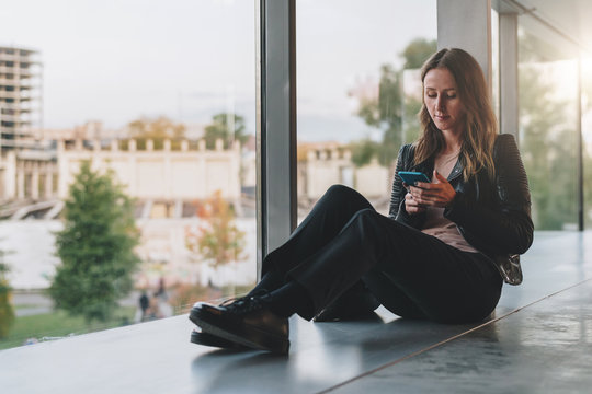 Young woman sits on floor near large window and uses smartphone.Hipster girl looking on screen of phone,blogging,chatting,browsing internet. Online marketing, education, distance work. Social network.