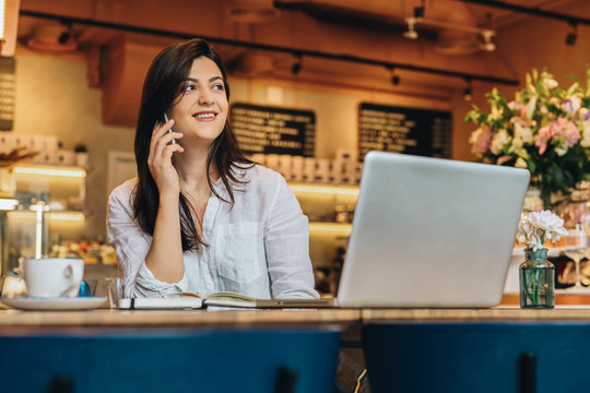 Young businesswoman dressed in white shirt is sitting in cafe at wooden table in front of laptop and talking on cell phone. Telephone conversations. Hipster girl is talking on phone with her friend.