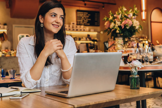 Young businesswoman wearing in shirt sitting in cafe at table, looking at window and smiling. On desk is laptop, notebook. Freelance,startup.Distance work,learning.Online marketing,education.Lifestyle