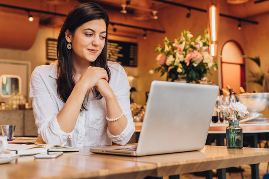 Young businesswoman wearing in shirt sitting in cafe at table, looking on screen of computer and smiling. Freelance, startup. Distance work, learning, blogging. Online marketing, education. Lifestyle.