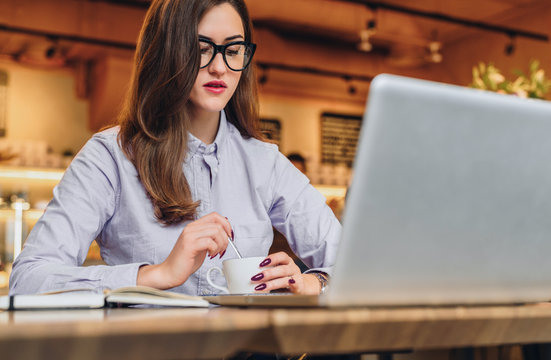 Young businesswoman wearing in shirt sitting in cafe at table and laughing. On desk is laptop, notebook, smartphone. Freelance, startup. Distance work, learning. Online marketing, education. Lifestyle