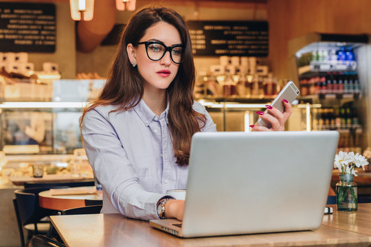 Front view. Young businesswoman is sitting in cafe at table, working on laptop. Hipster girl blogging, browsing internet, learning online, checking email. Online marketing, education, e-learning.