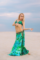 A beautiful blond girl in a golden with green dress with wings, suit is dancing an oriental, East dance in the desert on the sand. Beautiful exotic woman dancer in a costume for belly dancing.