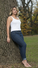Obraz premium Beautiful Caucasian female model poses in white tank top and blue jeans in park leaning against tree