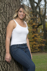Obraz premium Beautiful Caucasian female model poses in white tank top and blue jeans in park leaning against tree and smiling as looks down and right