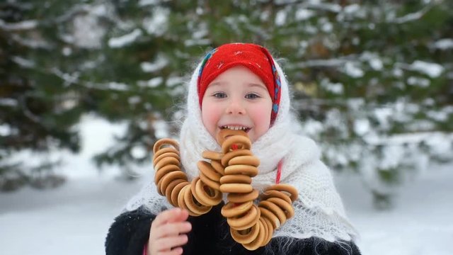 child girl in a fur coat and in a red scarf in Russian style eats bagels on a background of snow and forest. Celebration of carnival in Russia.