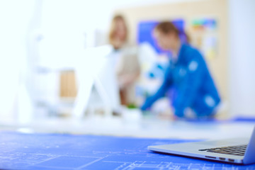 Two young woman standing near desk with instruments, plan and laptop.