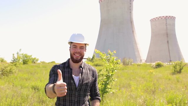 Portrait of a successful bearded male engineer in a white helmet against a background of cooling towers, he shows a thumbs-up.