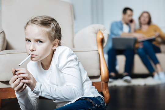 Cigarette. Poor blue-eyed little girl holding a cigarette and smoking while her parents working on their laptops