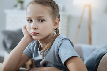 In the blues. Sad fair-haired blue-eyed little girl thinking and holding her head with her hand while sitting on the sofa