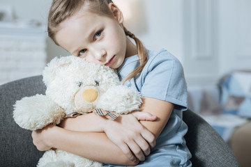 My bear. Miserable fair-haired blue-eyed little girl hugging and holding her teddy bear while...