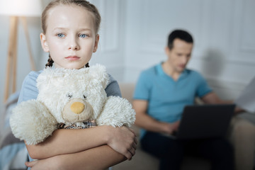 Bad mood. Unhappy blue-eyed fair-haired little girl holding her toy and standing in the room while her daddy working in the background