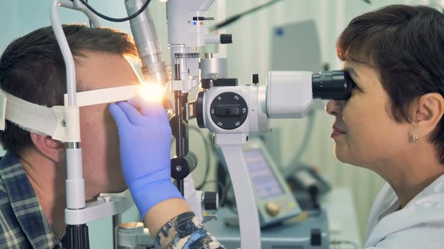 A female ophthalmologist checks young mans eyes with a slit lamp in a clinic. 4K.