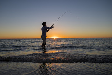 silhouette of fisherman with hat on the beach with fish rod standing on sea water fishing at sunset with beautiful orange sky in vacations