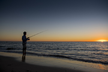 silhouette of fisherman with hat on the beach with fish rod standing on sea water fishing at sunset with beautiful orange sky in vacations