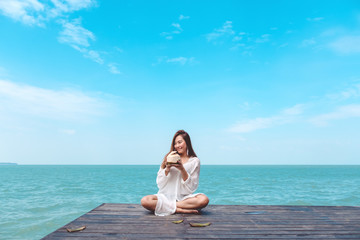 Fototapeta na wymiar A happy beautiful asian woman on white dress sitting at the wooden terrace and drinking coconut juice with sea and blue sky background