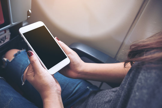 Mockup image of woman's hands holding a white smart phone with blank black desktop screen next to an airplane window