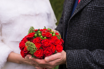 Winter wedding. Two wedding rings lies on a red wedding bouquet. Bride and groom holding red bouquet of fresh roses