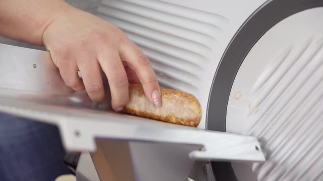 Electric food slicer. Meat slicing machine cutting processed meat products. Full aluminium alloy body and stainless steel blade. Closeup of female chef hand slices smoked sausage.