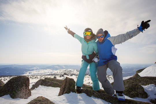 A guy and a girl snowboarders at the top of the mountain with their hands up, rejoice at the sun and the wind.