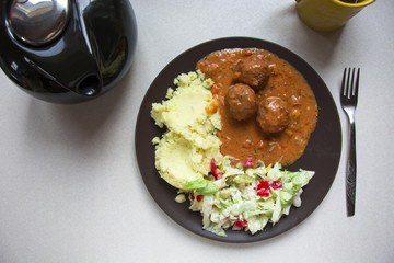 Meatballs in sauce with potatoes and salad on a plate, black kettle