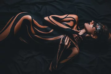  Mooi meisje met geschilderd lichaam en zwart masker in haar gezicht ligt op bed. LICHAAMSKUNST. © Stanislav