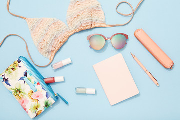 Beach holiday accessories, bikini, sunglasses, makeup bag, notebook on blue background, travel flat lay