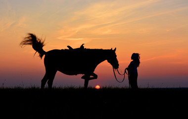 Romantic girl with horse on sunset or sunrise background. Beautiful sky, red setting sun. Rider and equine silhouette on horizon in summer evening.