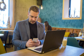 Handsome businessman is sitting in modern coffeehouse with smart phone and laptop and enjoying his morning coffee
