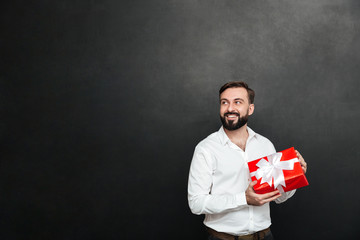 Picture of smiling bearded man holding red gift box with white ribbon and looking aside, over dark gray wall