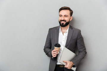 Portrait of cheerful male office worker posing on camera holding takeaway coffee and silver laptop,...