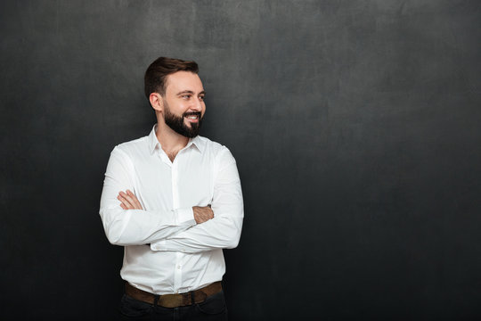 Portrait Of Positive Man In White Shirt Standing With Arms Folded, And Looking Away Over Dark Gray Background Copy Space