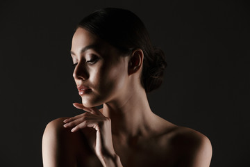 Naklejka premium Portrait of sensual beautiful woman looking aside while touching her chin in low lights, isolated over black background