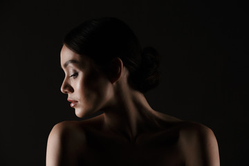 Beautiful portrait of half-naked elegant woman with dark hair in bun putting head aside, isolated over black background
