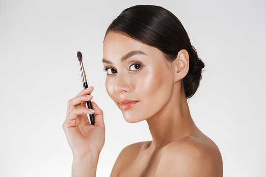 Half-turn picture of pleased woman with fresh skin looking on camera and holding make up brush for eyeshadow, isolated over white wall
