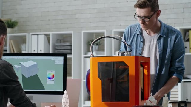 Tracking shot of young engineers in 3D printing studio: man and woman drafting 3D model on computer while their male colleague working with 3D printer