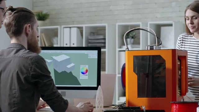 Tracking shot of young engineers working in team in 3D printing studio: two men designing 3D model on computer while woman controlling 3D printing process