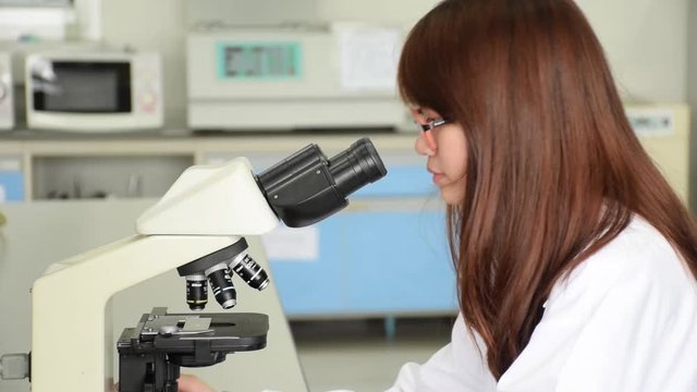 Closeup of Asian Thai university college student or scientist is looking through microscope eye piece and adjusting objective lens for microbiology test in science laboratory. 