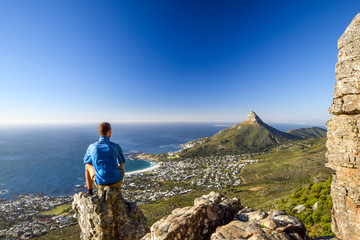 Fototapeta premium Young male hiker sitting on a rock at Kasteelspoort Hiking Trail in Table Mountain National Park in Cape Town, South Africa, facing the suburb of Camps Bay and Lion's Head mountain in the background.