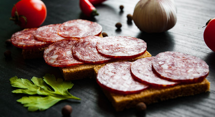 Tasty appetizer sandwich of thinly sliced spicy salami on bread on a dark wooden background