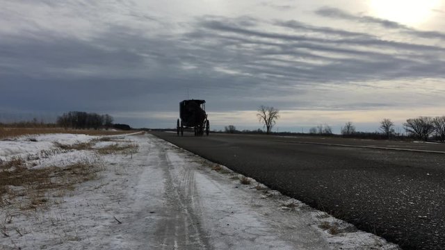 Amish buggy approaches camera on side of country road with the sounds of a horse gallop as it passes.