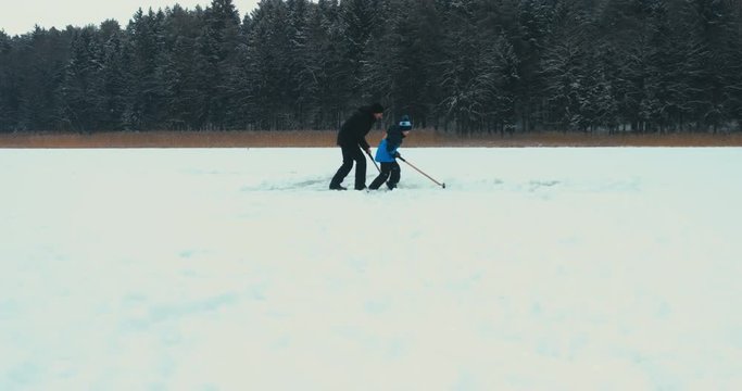 Father and son cleaning lake surface from snow before ice-skating and playing pond hockey. 4K UHD